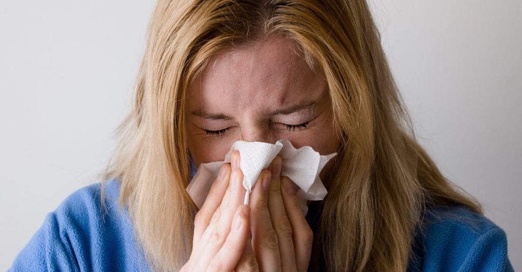 A woman in a blue robe is sneezing into a tissue, illustrating symptoms of a cold or flu indoors.